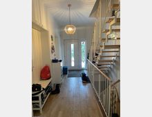 Heller Hausflur mit Holzoptik-Fliesenboden, weißer Eingangstür mit schmalen Glasfenstern und Deckenpendelleuchte. Rechts führt eine offene Treppe mit Holzstufen und weißem Geländer nach oben; links stehen eine Sitzbank und ein schmaler Schrank mit Garderobenbereich.