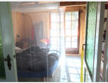 Ein einfach eingerichtetes Schlafzimmer mit Holzvertäfelung und einem großen Fenster, das Tageslicht hereinlässt. Im Zentrum steht ein Bett mit Moskitonetz, daneben ein kleiner Nachttisch mit Lampe, und an der Wand hängt Kleidung.