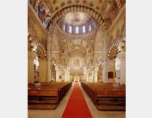 Innenansicht einer historischen Kirche mit kunstvoll verzierten Wänden und Decken. In der Mitte führt ein roter Teppich zwischen den hölzernen Kirchenbänken zum Altar, während Bögen und Säulen die Architektur prägen.