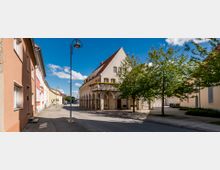 Straßenszene in einem historischen Stadtviertel mit einem Gebäude im traditionellen Stil, das Arkaden, einen Balkon mit Blumenkästen und ein steiles Satteldach besitzt. Entlang der Straße stehen Bäume, und der blaue Himmel ist mit einigen Wolken bedeckt.