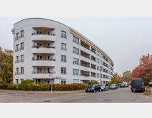 Wohngebäude mit weißer Fassade und mehreren Balkonen, das sich über vier Stockwerke erstreckt und an einer Straßenecke liegt. Umgeben von herbstlich gefärbten Bäumen, parkenden Autos und Gehwegen mit niedrigen Hecken.