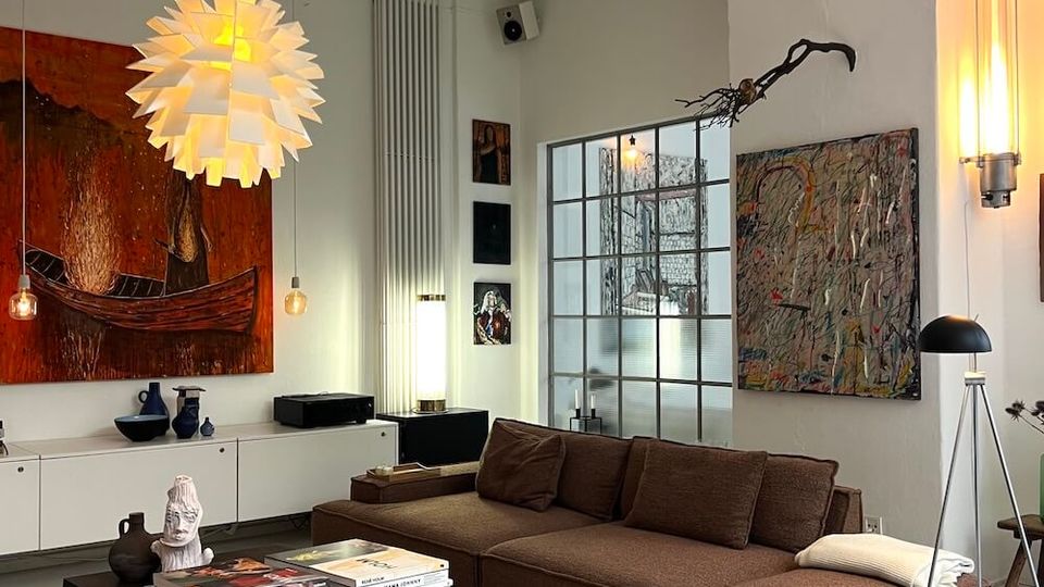 Modern living room with high ceilings and exposed beams, featuring a brown sectional sofa on a large rug and a black coffee table stacked with books. White walls display large abstract artwork, a sculptural pendant light hangs overhead, and a gridded glass door and tall radiator sit along the back wall.