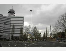 Straßenszene mit einem modernen Bürogebäude links, das den Schriftzug „rbb“ trägt, und dem Funkturm im Hintergrund rechts. Die Straße ist von kahlen Bäumen gesäumt, und Ampeln und Verkehrsschilder sind sichtbar.