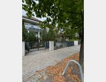 Mehrfamilienhaus mit einer Fassade in hellen Farben und einem Balkon mit Glasgeländer im Hintergrund. Im Vordergrund ein Gehweg aus Pflastersteinen, Laub auf dem Boden und ein schmiedeeisernes Tor mit Hecken und Bäumen drumherum.