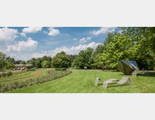 Eine Parklandschaft mit einer großen Wiese, bepflanzten Blumenbeeten und geschwungenem Weg. Auf der Wiese stehen moderne Metallskulpturen, darunter eine abstrakte geometrische Figur, umgeben von Bäumen und vereinzelten Besuchern.