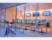 Ein moderner Raum mit mehreren Computer-Arbeitsplätzen, die in einer Reihe hinter einer Glaswand angeordnet sind, mit Monitoren, Tastaturen und Telefonen. Im Vordergrund steht ein stationäres Trainingsfahrrad, während Monitore und Bildschirme an den Wänden hängen, die Informationen anzeigen.