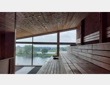Innenansicht einer Sauna mit Holzbänken und Holzdecke, durch ein großes Panoramafenster ist ein Fluss, Bäume und eine Brücke in einer ländlichen Umgebung zu sehen.