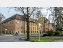Gelbes, historisches Gebäude mit mehreren großen Fenstern und einem Satteldach, umgeben von Bäumen. Im Vordergrund eine Kreuzung mit Straßenschildern, einem Fahrradfahrer und wenigen Grünflächen.