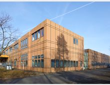 Flaches Büro- oder Industriegebäude mit beigefarbener Fassade und rechteckigen Fenstern, umgeben von kahlen Bäumen und einer asphaltierten Straße. Der Himmel ist hellblau, und Schatten der Bäume fallen auf die Fassade.