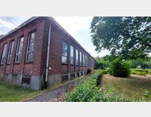 Backsteinbau mit großen rechteckigen Fenstern in einer ländlichen Grünanlage. Ein schmaler Gehweg führt entlang des Gebäudes, umgeben von Sträuchern und Bäumen.