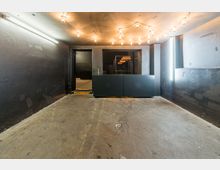 Ein einfacher Raum mit schwarzem Wänden und Betonboden, beleuchtet von hängenden Glühbirnen und LED-Leuchten an der Wand. Im Hintergrund befindet sich eine schwarze Theke vor einer Türöffnung, die zu einem weiteren Raum mit Kleiderständern führt.