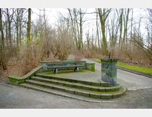 Sitzbank und Mülltonne auf einer kleinen gepflasterten Plattform mit Stufen in einem winterlichen Park. Umgeben von laubfreien Bäumen und Sträuchern, mit angrenzenden Gehwegen im Hintergrund.