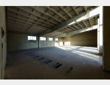 Großer leerer Raum mit Betonwänden und -boden, sichtbarem Betondach mit Balkenstruktur und mehreren rechteckigen Fensteröffnungen, die Tageslicht einlassen. Im Boden befinden sich vertiefte rechteckige Bereiche, und eine einzelne Person steht im Hintergrund im Licht.