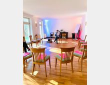 Ein heller Seminarraum mit Holzfußboden und Stuhlkreis, auf den Stühlen liegen pinkfarbene Mappen. Im Hintergrund stehen eine Person mit Flipchart, ein Mann, der ein Blatt liest, und ein Tisch mit einer Blumenvase. Natürliches Licht fällt durch große Fenster.