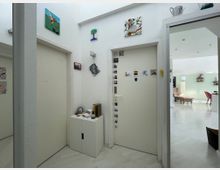 Ein heller Eingangsbereich mit weißen Wänden, einer Tür verziert mit Fotos und Stickern, und einer kleinen Kommode mit Dekorationsgegenständen. Rechts führt der Raum in einen offenen Wohnbereich mit mintfarbenen Wänden, rosa Stühlen und dekorativen Regalen.