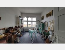Ein Arbeitszimmer mit passenden historischen Werkzeugen und Haushaltsgegenständen. Der Raum hat große, dekorative Fenster mit Ausblick auf eine Siedlung, weiße Wände und eine Leiter sowie einen Tisch mit Geschirr in der Mitte.