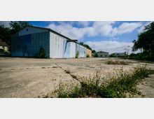 Industriegelände mit verlassenen Lagerhallen aus Wellblech und Betonboden, umgeben von wildem Gras und Büschen. Im Hintergrund sind weitere Gebäude und Bäume zu sehen, der Himmel ist teilweise bewölkt.