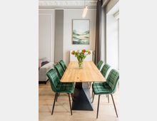 Moderner Essbereich mit langem Holztisch und sechs grünen, gepolsterten Stühlen auf hellem Holzboden. An der Wand hängen ein Landschaftsbild über einem Heizkörper; daneben ein großes Fenster mit dunklen Vorhängen und eine schlichte Pendelleuchte über dem Tisch.