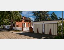 Backsteinhaus mit weißem Zaun in einer ruhigen, gepflasterten Straße, daneben weiße Garagen mit braunen Toren. Im Hintergrund Bäume und ein klarer blauer Himmel.