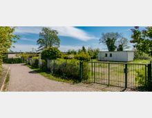 Ein gepflasterter Weg führt an einem umzäunten Grundstück mit Grünflächen und niedriger Vegetation vorbei. Im Hintergrund steht ein weißes, flaches Gebäude mit zwei kleinen Fenstern, umgeben von Bäumen und Sträuchern unter einem blauen Himmel.