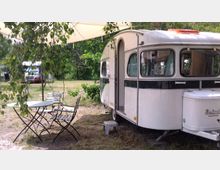 Ein weißer Wohnwagen mit schwarzen Akzenten steht auf einer grasbedeckten Fläche, umgeben von Bäumen. Im Vordergrund befindet sich ein kleiner Tisch mit zwei Metallstühlen, teils beschattet von einem Sonnensegel und herabhängenden Ästen.