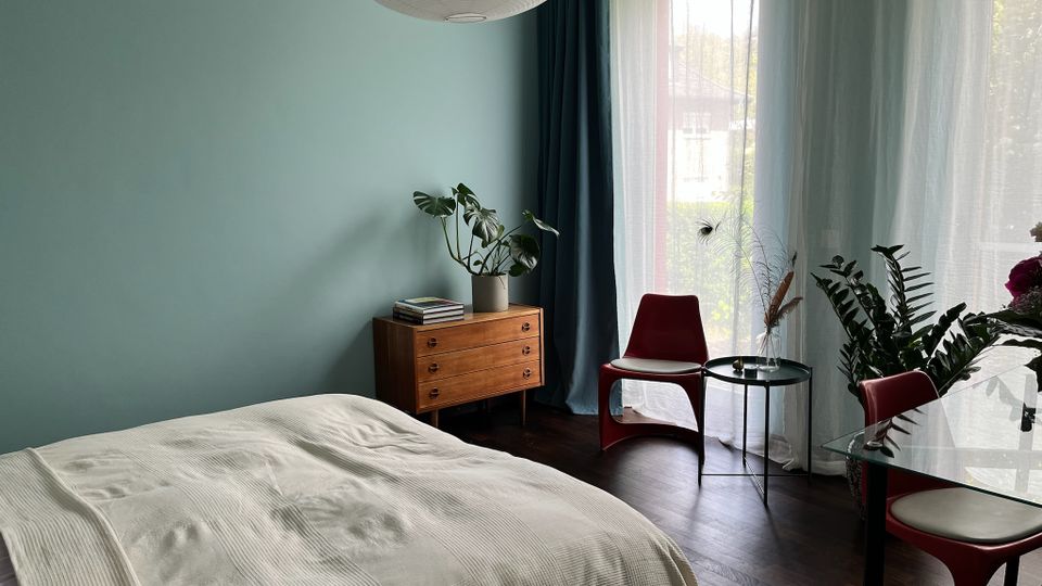 Helles Schlafzimmer mit mintfarbener Wand, großem Fenster mit transparenten Vorhängen und dunklem Holzboden. Neben dem Bett stehen eine Holzkommode mit Pflanze, ein kleiner runder Beistelltisch und rote Stühle an einem Glastisch.
