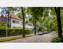 Eine gepflasterte Wohnstraße mit Bäumen gesäumt, auf der linke Seite befinden sich weiße Einfamilienhäuser mit roten Satteldächern und Vorgärten. Rechts parken mehrere Autos am Straßenrand, während die Umgebung dicht mit Grün bewachsen ist.