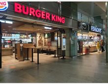 Eingangsbereich eines Burger King-Restaurants und eines benachbarten Fish-&-Chips-Ladens in einem modernen, hell beleuchteten Einkaufszentrum. Der Burger King ist mit Selbstbedienungsterminals, Holzsitzgelegenheiten und Leuchtschrift ausgestattet, während der Fish-&-Chips-Laden eine Verkaufstheke mit Auslage zeigt.