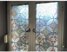 Zwei Fenster mit buntem, ornamentverziertem Milchglas, das geometrische und florale Muster zeigt. Der Fensterrahmen ist aus hellem Holz mit schwarzen Griffen, die sich in der Mitte treffen.