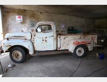 Alter Pick-up-Truck in einer Werkstatt mit rostigen Stellen auf der Karosserie. Im Hintergrund sind verblasste Wandschilder, Werkzeuge und ein weiteres altes Fahrzeug zu sehen.