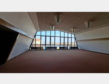 Großer, leerer Innenraum mit schrägen Wänden, Teppichboden und abgehängter Decke mit mehreren Deckenleuchten. Eine breite Fensterfront über die gesamte Stirnseite bietet Ausblick auf Gebäude und eine offene Außenfläche.