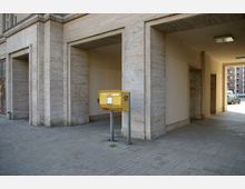 Gelber Briefkasten der Deutschen Post vor einem Gebäudeeingang mit hellen Steinwänden und drei großen Durchgängen. Der Boden besteht aus grauen Pflastersteinen, und im Hintergrund sind ein Auto und Teile einer Straße mit Wohngebäuden sichtbar.