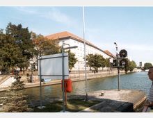 Ein Fluss oder Kanal mit einem modernen Gebäude mit rotem Dach im Hintergrund. Am Ufer stehen Bäume und Schilder, im Vordergrund befindet sich eine kleine Plattform mit einer Boje und einer Signalvorrichtung.