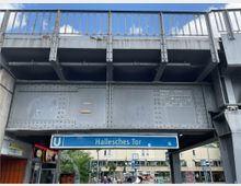 U-Bahnhof "Hallesches Tor" mit einer stählernen Brückenkonstruktion darüber, die mit Nieten verstärkt ist. Darunter befindet sich das blaue U-Bahn-Schild, seitlich sind Geschäfte und dahinter eine belebte Straßenkreuzung mit Gebäuden und Bäumen sichtbar.