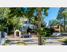 Wohnstraße mit Einfamilienhäusern, umgeben von Bäumen und Grünflächen. Im Vordergrund führt eine gepflasterte Straße entlang, mit geparkten Autos und einer Kreuzung, die von Straßenschildern und einem Baum gesäumt ist.