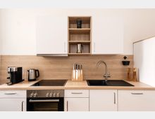 Moderne Küchenzeile mit weißen Schränken, einer Holzarbeitsplatte und einer Rückwand in Holzoptik. Zu den sichtbaren Elementen gehören ein Elektroherd, ein Wasserkocher, ein Kaffeemaschine, ein Spülbecken mit Armatur, ein Messerblock sowie offene Regale mit Kaffekannen.