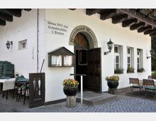 Fassade eines historischen Gasthauses mit der Aufschrift „Wirtshaus Moorlake“, gebaut 1840. Vor dem Gebäude befinden sich ein Eingang mit Holztür, Blumenkübel, Laternenlampen, mehrere Fenster mit Blumenkästen sowie outdoor Sitzmöglichkeiten mit gelb-weiß karierten Tischdecken.