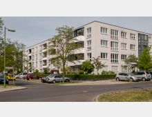 Wohnhaus in einem urbanen Viertel mit vier Etagen und hellgrauer Fassade, großen Fenstern und Balkonen. Vor dem Gebäude befinden sich Bäume, geparkte Autos und eine asphaltierte Straße mit Gehwegen.