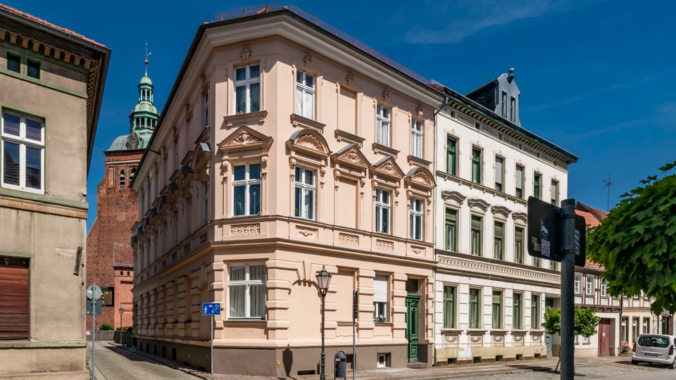 Straßenecke in einer Altstadt mit dreigeschossigen, reich verzierten Gründerzeit-Häusern in Pastellfarben, großen Fenstern und Stuckfassaden. Im Hintergrund ragt ein Backsteinturm mit grünem Kupferdach auf; am Straßenrand stehen Laterne, Verkehrszeichen und ein Baum.