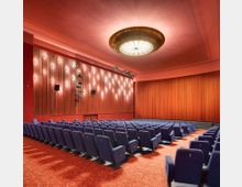 Kinosaal mit rot gepolstertem Teppichboden, blauen Sesseln in Reihen angeordnet, einer großen Leinwand hinter einem geschlossenen roten Vorhang und einer seitlichen Holzwand mit integrierter Beleuchtung. An der Decke hängt eine runde, zentral platzierte Lichtinstallation.