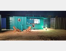 Ein umgebauter türkisfarbener Schiffscontainer mit einer geöffneten Verkaufsfläche, beleuchtet in einer sandigen Außenumgebung; davor stehen zwei klappbare Liegestühle und ein Stehtisch. Im Hintergrund sind weitere Container zu sehen.