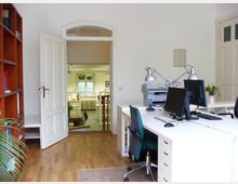 Helles Büro mit Holzboden, weißen Wänden und weißen Möbeln. Im Vordergrund stehen Schreibtische mit Computern und Lampen, dahinter eine offene Tür, die in einen weiteren Raum mit großen Fenstern und Tageslicht führt.