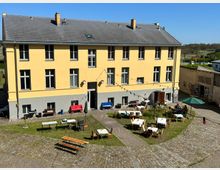 Gelbes zweistöckiges Gebäude mit grauem Sockel und mehreren Fenstern, umgeben von einem gepflasterten Hof. Im Hof stehen Tische, Bänke und Stühle, teilweise mit Tischdecken, sowie wenige Dekorationen wie bunte Lichterketten; im Hintergrund ist ländliche Umgebung sichtbar.