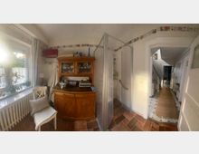 Helles Badezimmer mit gemustertem Fliesenboden, einer Glasdusche auf der rechten Seite und einem Holzschrank mit Glasvitrine links daneben. Eine Sitzgelegenheit und dekorative Pflanzen stehen vor einem Fenster mit weißem Vorhang, das seitlich Licht hineinlässt, und ein angrenzender Flur ist im Hintergrund sichtbar.