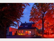Ein kleines zweistöckiges Haus mit gelber Fassade, beleuchtet durch rotes und warmweißes Licht, steht inmitten von großen Bäumen mit rot angestrahltem Laub, vor einem abendlichen, blauen Himmel. Im Vordergrund befindet sich ein gepflasterter Hof, rechts und links von Bäumen eingerahmt.