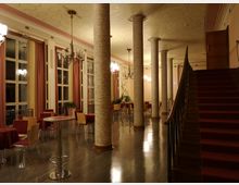 Ein eleganter Innenraum mit klassischen Säulen, einer Holztreppe mit rotem Teppich, hohen Fenstern mit Vorhängen und mehreren Tischen mit roten Tischdecken und Stühlen. Die Decke ist mit dekorativen Mustern gestaltet, und Kronleuchter sorgen für Beleuchtung.