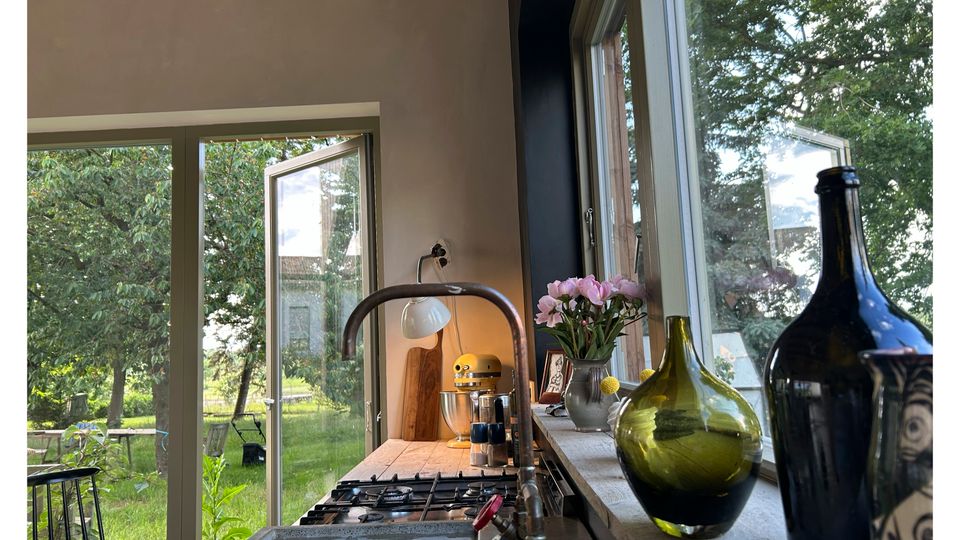 Helle Küche mit großer Arbeitsplatte, Gasherd und Wasserhahn vor bodentiefen Fenstern. Auf der Fensterbank stehen Glasflaschen und eine Vase mit Blumen; draußen ist ein grüner Garten mit Bäumen zu sehen.