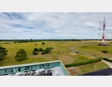 Weite Wiesenlandschaft mit vereinzelten Bäumen und Sträuchern, im Hintergrund ein Waldstreifen. Rechts im Bild steht ein großer, rot-weißer Funkturm, daneben verlaufen ein asphaltierter Weg und einige parkende Fahrzeuge.