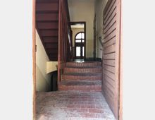 Treppenhaus in einem Altbau mit roten Ziegelstufen, einer hölzernen Treppe und wandseitigen Holzpaneelen. Im Hintergrund ist eine gläserne Doppeltür mit einem abgerundeten oberen Fenster zu sehen.