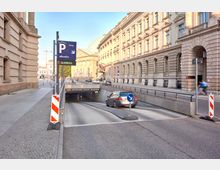 Einfahrt zu einem öffentlichen Parkhaus unter einer belebten Straße in einer Stadt mit klassischer Architektur. Auf der rechten Seite sind historische Gebäude mit Steinfassaden, und links ein breiter Gehweg entlang der Häuser.